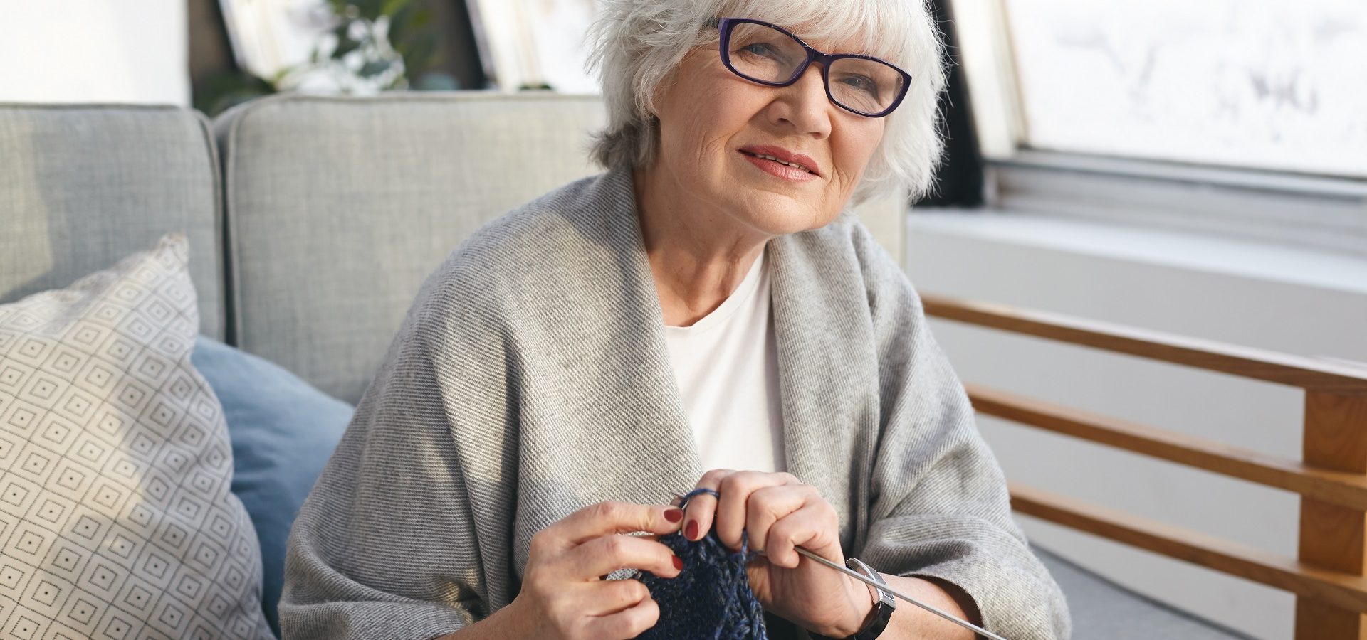 Picture of neat retired woman wearing wide scarf and eyeglasses knitting warm jumper for her daughter. Attractive senior female knitter working from home, handmaking winter clothes for sale